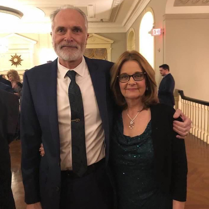 A man and woman are smiling towards the camera. He is wearing a black suit and tie.