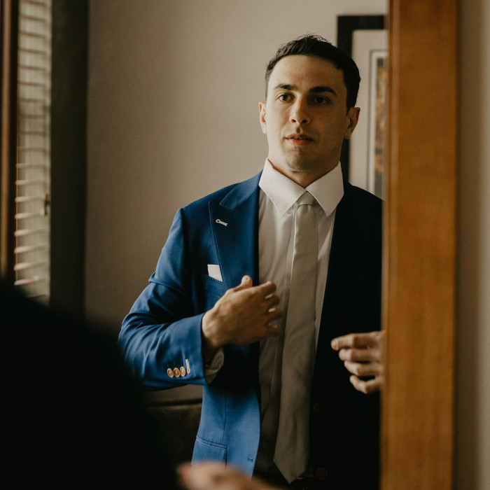 A man is wearing a suit and looking at his reflection in a large mirror