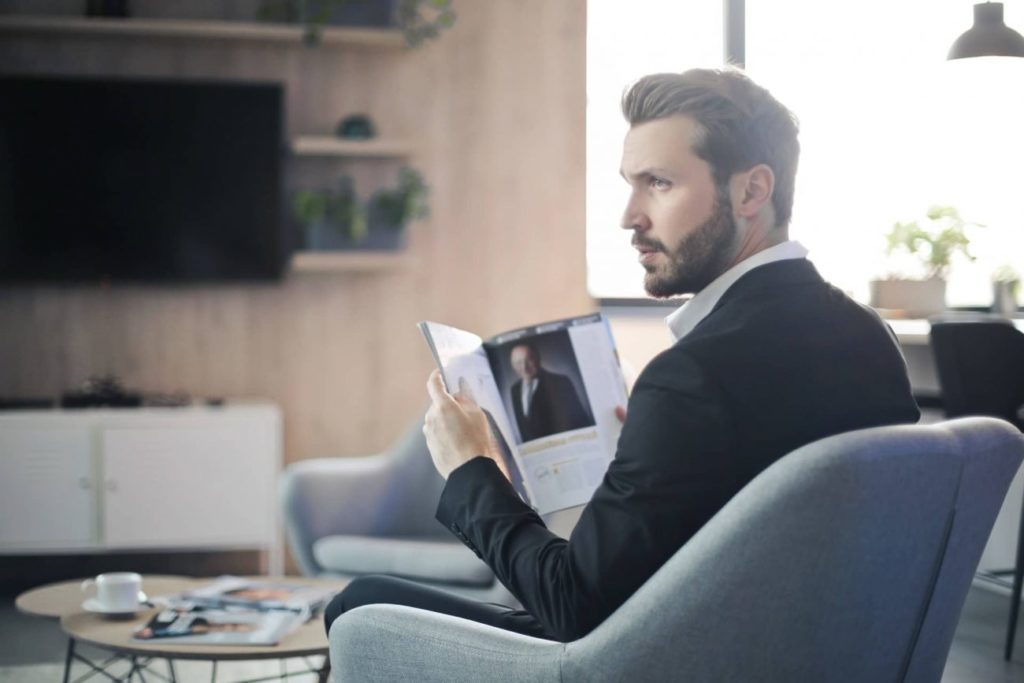 Man with well groomed beard and trimmed sideburnssitting on chair holding a magazine