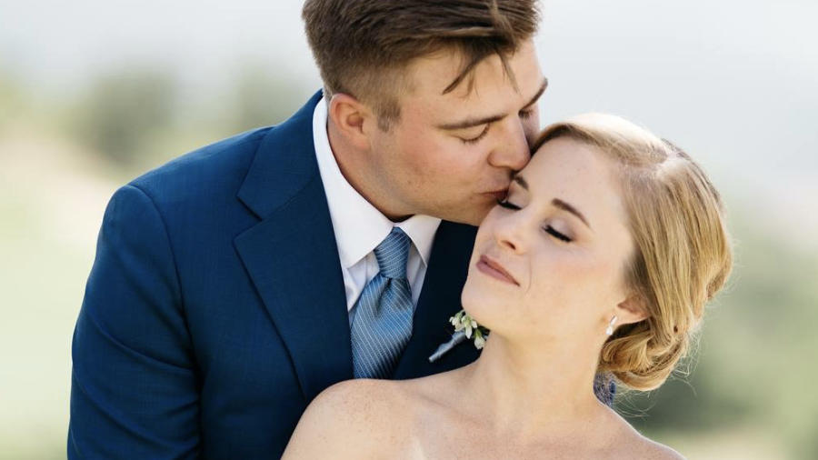 A married couple is posing. The man is kissing the woman on the head.